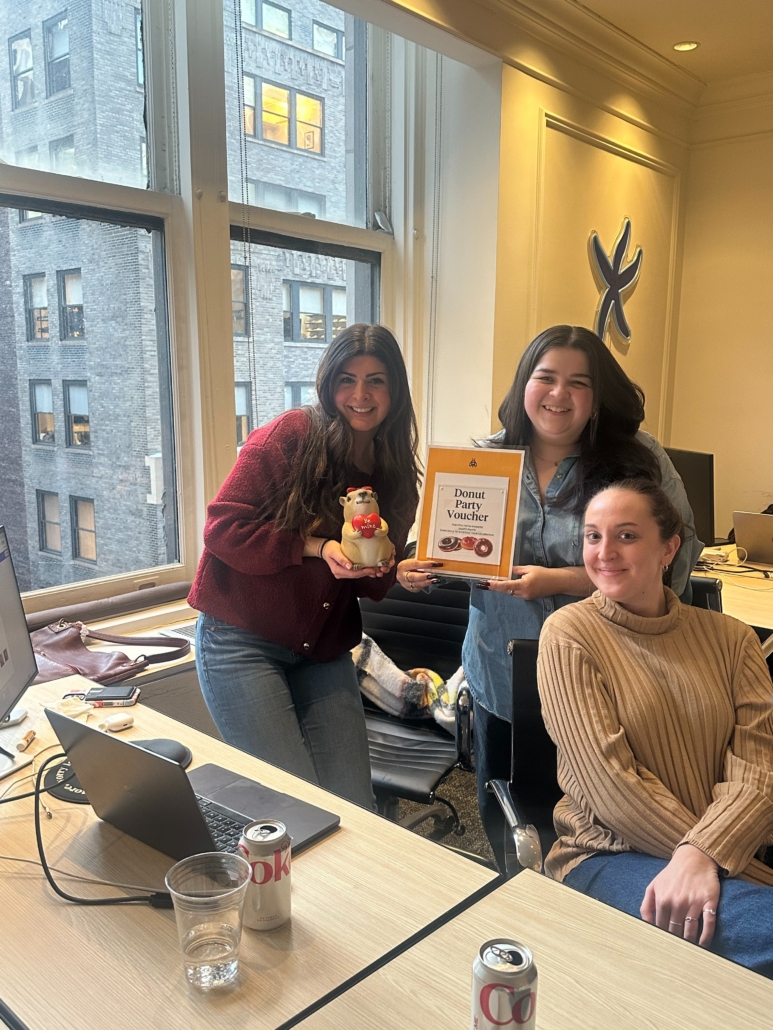 Three women in an office pose for a photo, one holds an orange Donut Party Voucher while another holds a small bear figurine, laptops and drinks on the desk behind them.