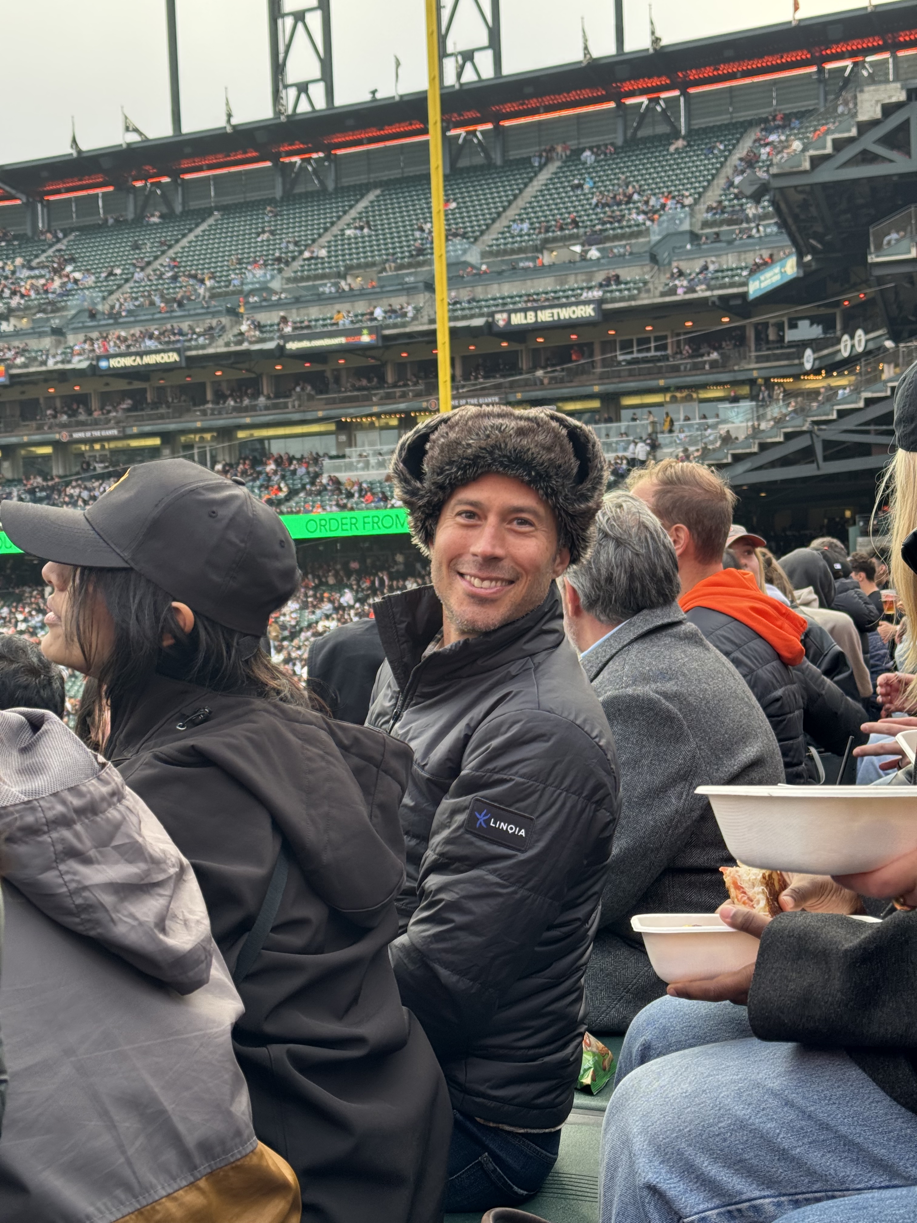 Smiling man in a black fur-lined hat at a crowded stadium.