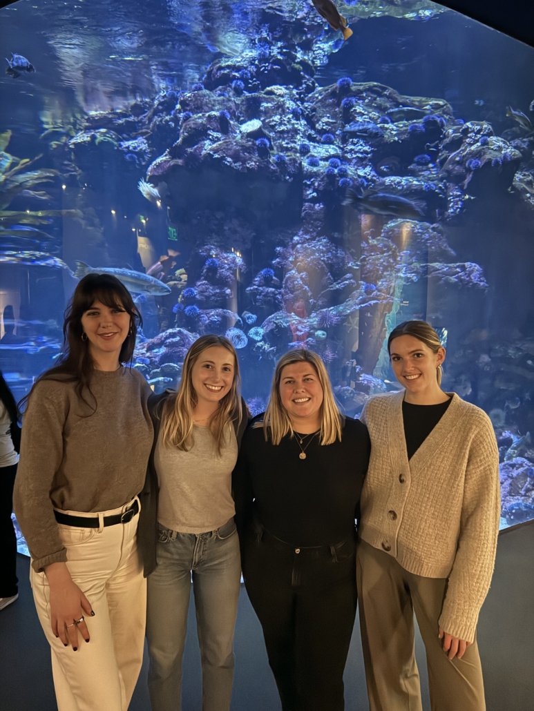 Four women posing together in front of a large aquarium tank with coral and fish in the background.