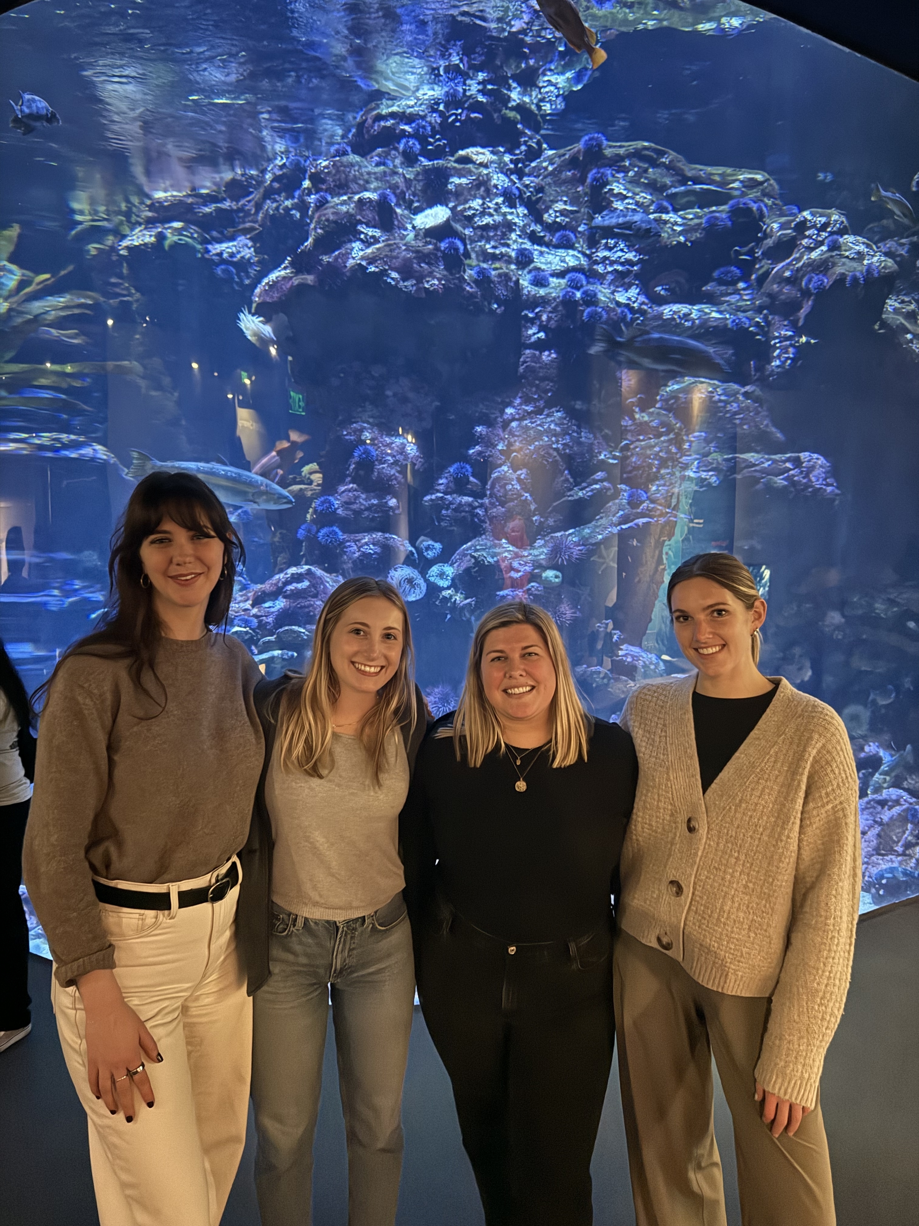 Four women posing together in front of a large aquarium tank with coral and fish in the background.
