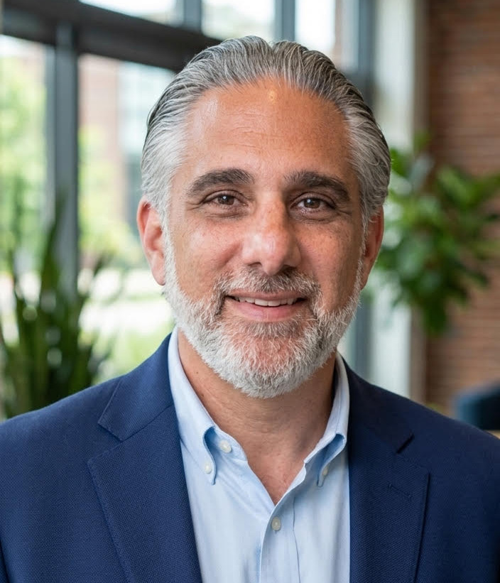Portrait of a smiling mature man with gray hair and beard, wearing a blue blazer and light blue shirt, indoors near large windows.